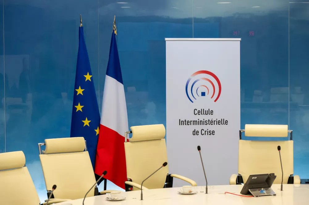 Salle de la cellule interministérielle de crise : fauteuils vides, micros, drapeaux français et européen, et kakémono au logo de la CIC