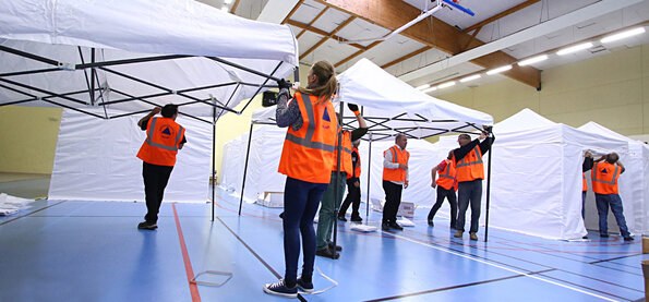 Exercice de crise dans un gymnase : des participants en gilet orange montent des tentes blanches pour un centre d'accueil