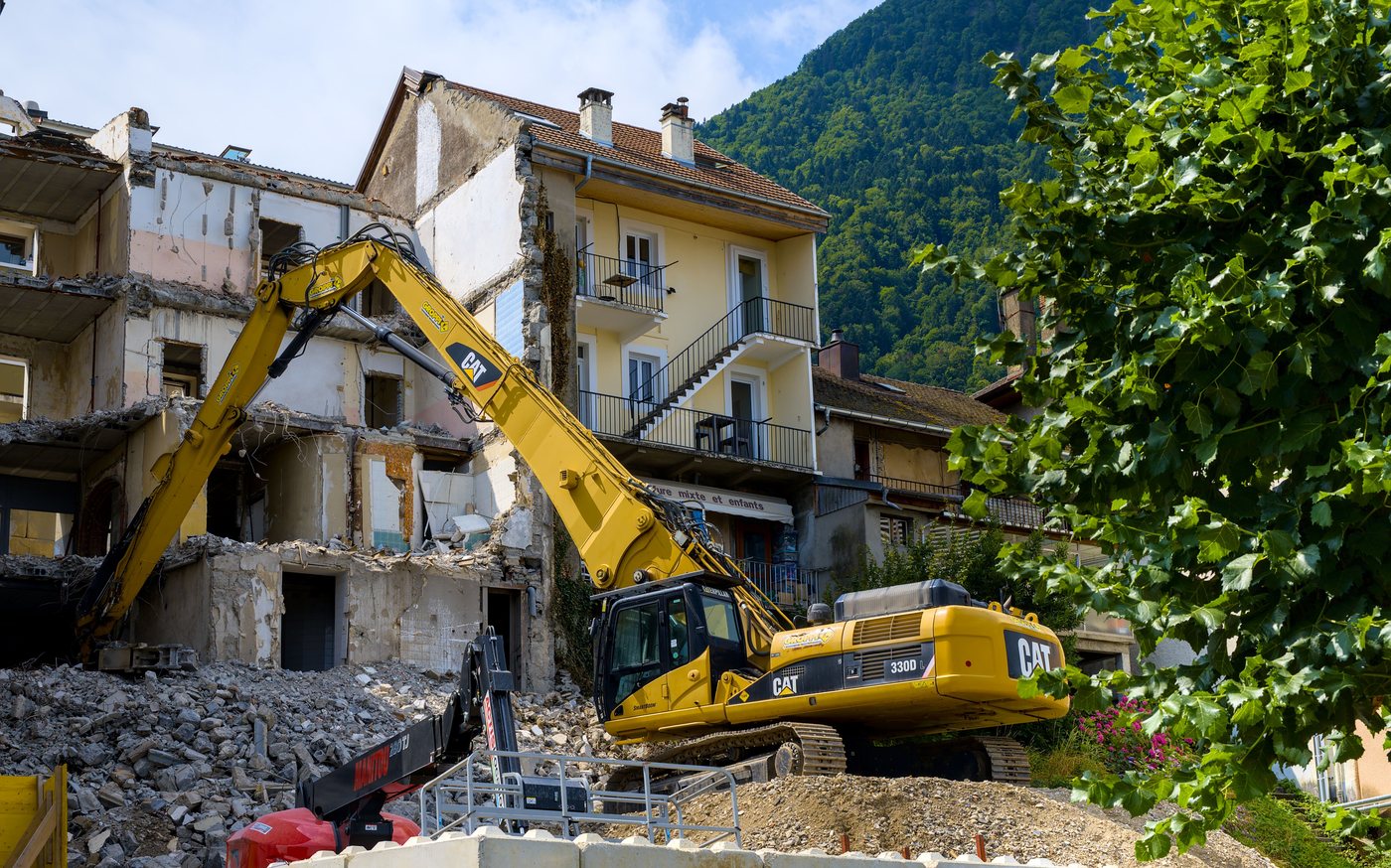 Pelle mécanique démolissant un immeuble mitoyen d'un bâtiment resté intact, dans un bourg de montagne