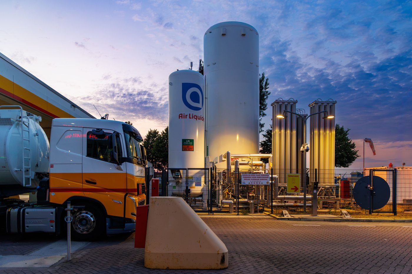 Station de ravitaillement au crépuscule : camion-citerne, réservoir cryogénique et vaporiseurs derrière une clôture grillagée