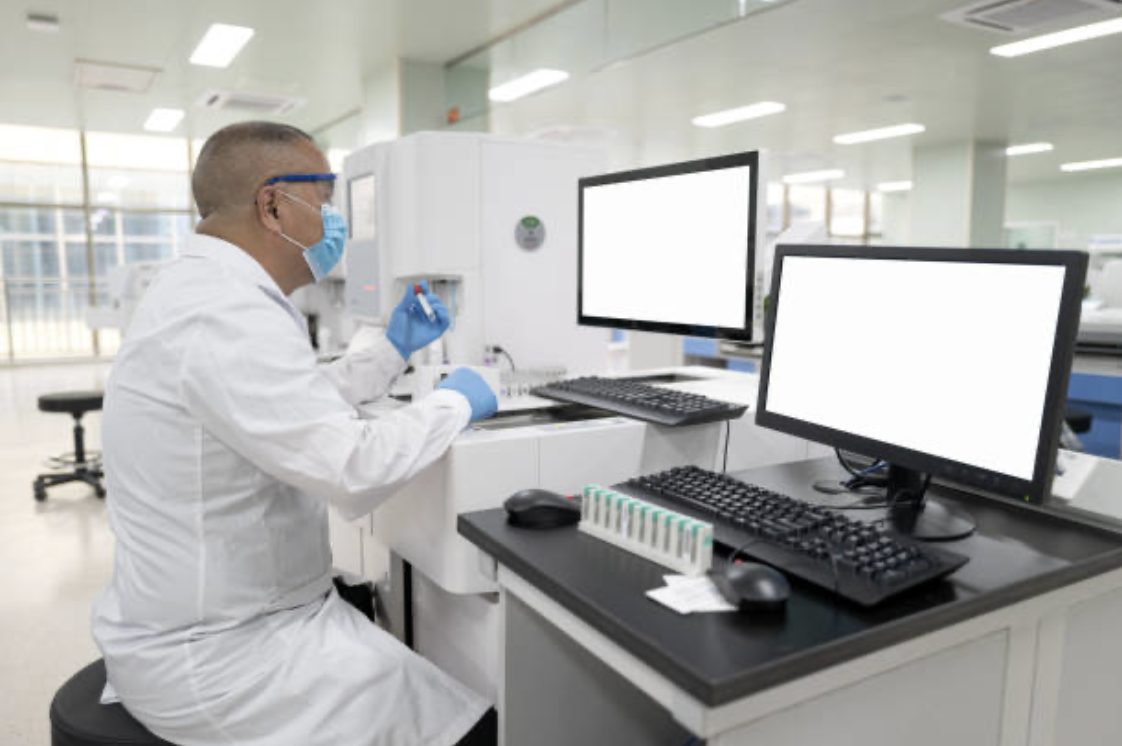Un technicien de laboratoire manipule des tubes devant deux écrans vides, dans un plateau technique d'établissement de santé