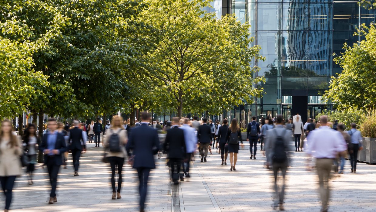 Flux de personnes en tenue de travail sur une esplanade arborée, devant une façade de verre